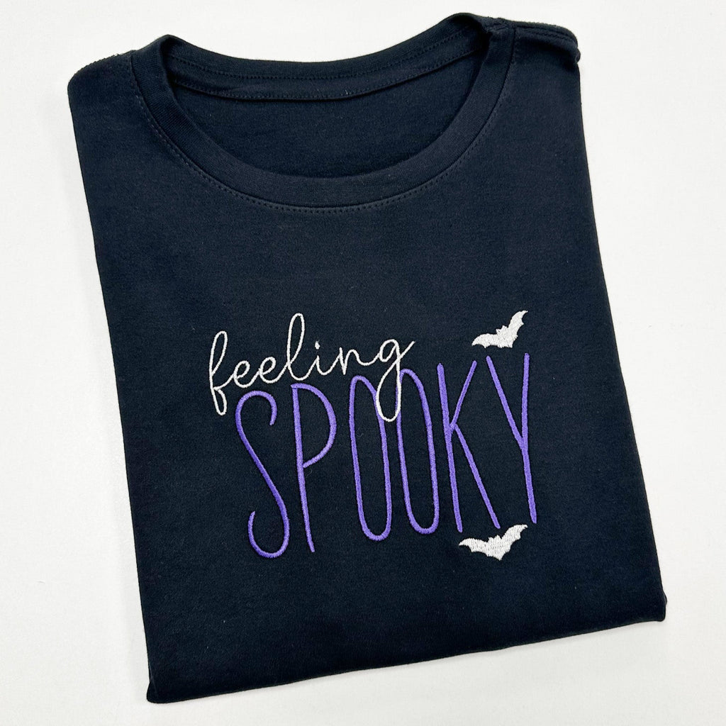 Folded black t-shirt with 'feeling spooky' text and bat graphics on a white background
