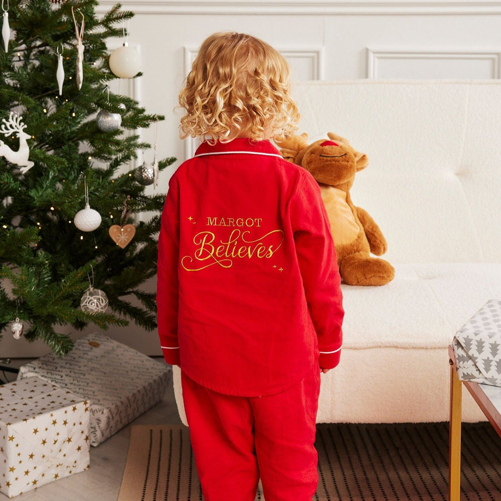 A child standing in a room with a red Christmas tree and presents, wearing traditional red pyjamas with 'MAGICAL BELIEFS' embroidered on the back.